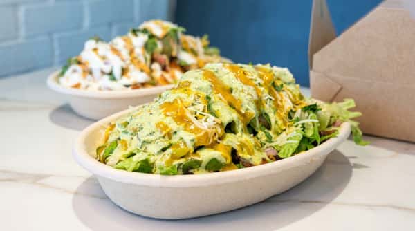 Two burrito bowls with greens, rice, cheese, and various sauces in eco-friendly containers.