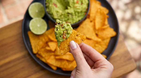Hand holding a tortilla chip with guacamole, blurry bowl of guacamole and lime halves in background.