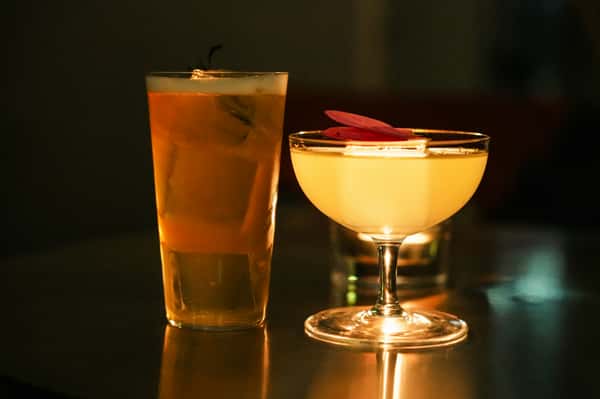Tall iced cocktail beside a coupe glass with a yellow drink garnished with pink flower petals, softly lit in a dark setting.