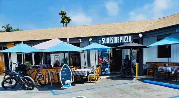 Outdoor patio seating at Surfside Pizza with blue umbrellas, checkered tablecloths, and surf-themed decor, showcasing a relaxed coastal pizzeria atmosphere.