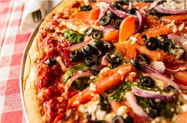 Close-up of a freshly baked vegetarian pizza topped with black olives, red onions, spinach, tomatoes, and crumbled cheese on a golden crust in a cozy Italian restaurant setting.