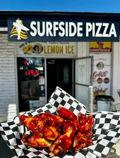 Basket of saucy barbecue chicken wings in checkered paper held outside Surfside Pizza, highlighting crispy wings and bold flavor at a local Italian restaurant.