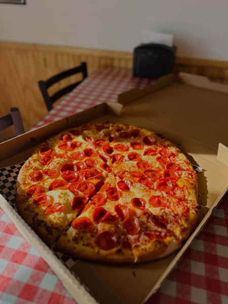 Pepperoni pizza in an open takeout box on a red and white checkered tablecloth, featuring a golden crust, melted mozzarella, and crispy pepperoni slices in a cozy pizzeria setting.