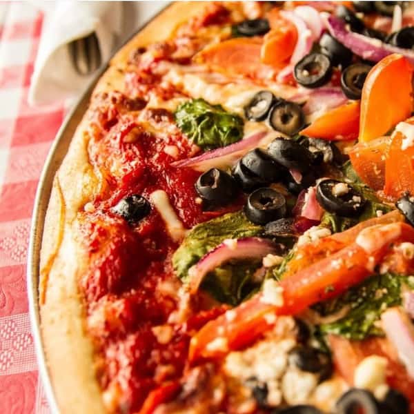 Close-up of a freshly baked vegetarian pizza topped with black olives, red onions, spinach, tomatoes, and crumbled feta over rich tomato sauce and melted mozzarella, served on a checkered tablecloth in a cozy Italian restaurant setting.