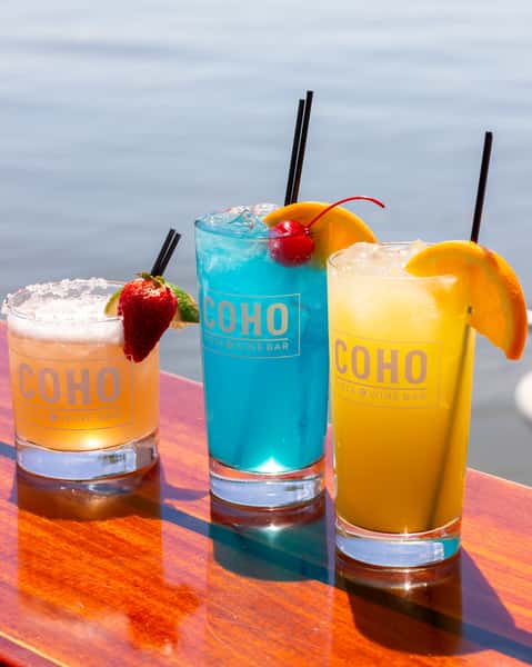 Three colorful cocktails in COHO glasses on a table by the water