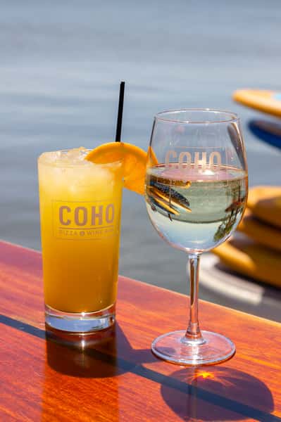 Two COHO cocktails, an orange drink and a glass of white wine, on a table by the water