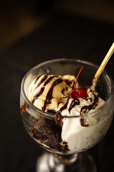 A decadent glass with our hot fudge sundae, with vanilla ice cream, whipped cream, brownie bits, chocolate drizzle and a cherry on top.