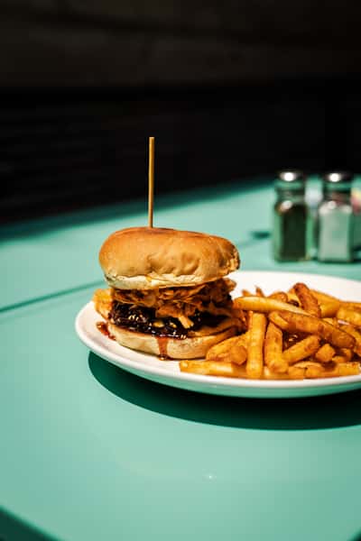 The Korean BBQ. Beef patty, kimchi coleslaw, chili garlic aioli, Korean BBQ sauce, toasted sesame seeds on a bun. Golden fries six next to the burger on a white plate, with a turquoise table underneath with salt and pepper shakers in the background.