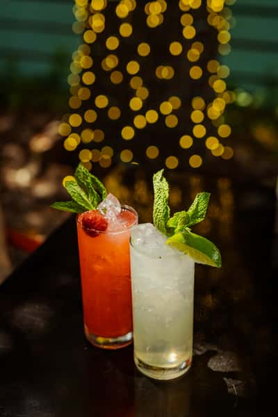 our raspberry mojito and classic mojito sit in tall glasses, with mint sitting on top. They are on a black bar with twinkle lights in the background.