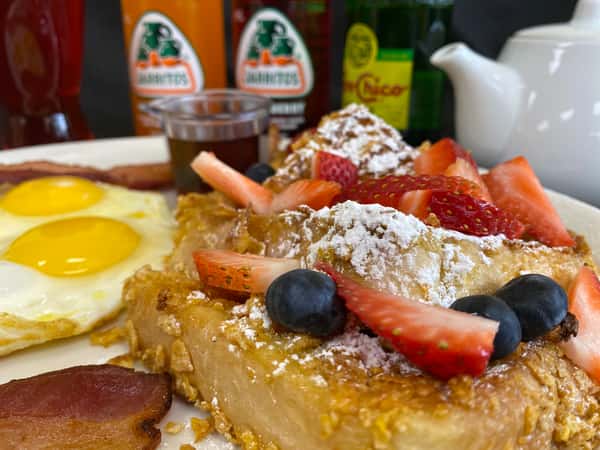 A close-up of a sweet and savory breakfast from Boulevard Cafe, featuring crispy-coated French toast topped with strawberries, blueberries, and powdered sugar, alongside sunny-side-up eggs and bacon.