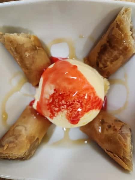 Thai dessert featuring crispy fried banana spring rolls served with a scoop of vanilla ice cream topped with strawberry syrup and a drizzle of honey.