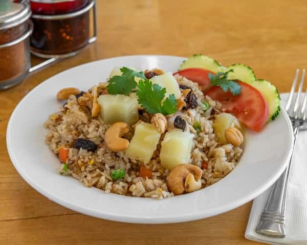 Pineapple fried rice topped with cashews, raisins, and pineapple chunks, garnished with fresh cilantro, sliced tomato, and cucumber—classic sweet and savory Thai rice dish.