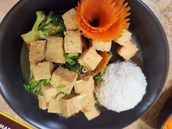 Bowl of crispy tofu cubes stir-fried with mixed vegetables, including broccoli and carrots, accompanied by a mound of white rice and a decorative carrot flower—perfect for a vibrant vegan meal.
