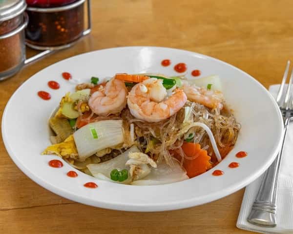 Stir-fried glass noodles with shrimp, egg, onions, and mixed vegetables, garnished with sriracha dots around the plate—classic Thai-style pad woon sen served in a white bowl with utensils.