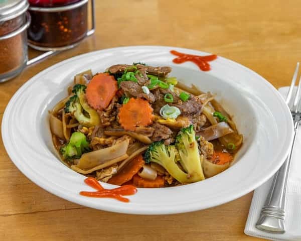 Stir-fried Thai drunken noodles (Pad Kee Mao) with wide rice noodles, beef, broccoli, carrots, and egg in a spicy basil soy sauce, garnished with scallions and chili sauce.