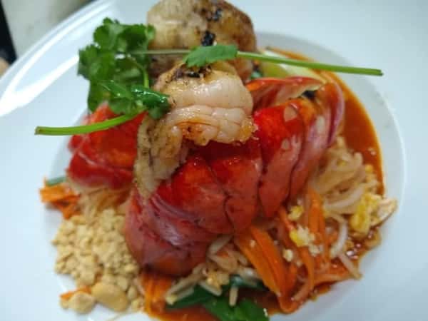Elegant Thai lobster Pad Thai featuring a split lobster tail and grilled shrimp atop stir-fried rice noodles with egg, bean sprouts, carrots, crushed peanuts, and fresh herbs, all coated in a rich tamarind sauce—served on a white plate for a gourmet Thai seafood experience.
