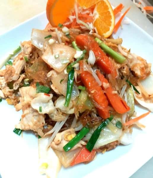 Savory Thai stir-fried glass noodles with chicken, carrots, onions, and bean sprouts, topped with scallions and orange slices—served on a square white plate for a vibrant presentation.