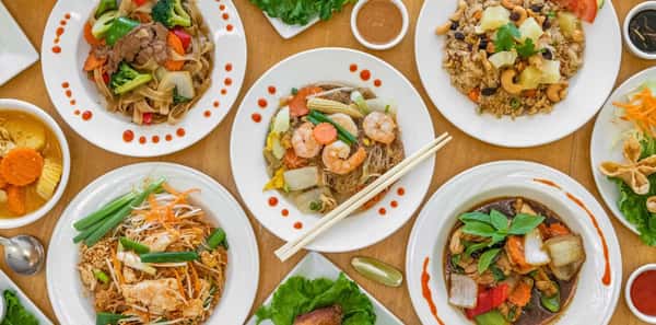 Top-down view of a colorful Thai food spread featuring dishes like Pad Thai, stir-fried glass noodles with shrimp, pineapple fried rice with cashews, red curry with tofu, and Thai basil stir-fry. Each plate is beautifully garnished with fresh vegetables, herbs, and chili sauce, showcasing authentic Thai cuisine on a wooden table.