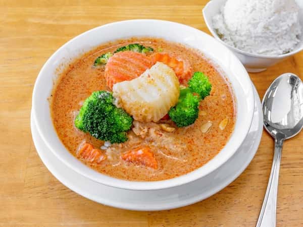 Creamy Thai red curry with scallops, broccoli, and carrots, served in a white bowl with a side of jasmine rice, showcasing a rich and spicy seafood entrée.