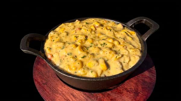 Creamy corn dip with visible corn kernels, herbs, and red flecks, in a black cast iron dish on a wooden board.