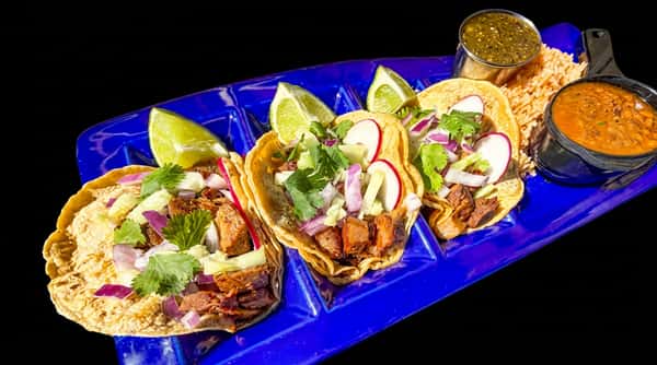 Three street tacos with meat, cilantro, red onion, radishes, and lime wedges on a blue tray, with rice and beans.