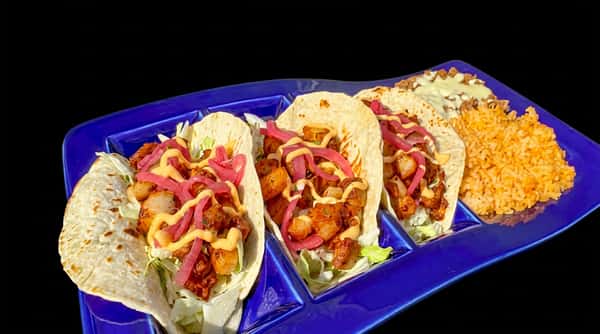 Three tacos with fried shrimp, shredded lettuce, pickled red onions, and sauce, served with rice and beans on a blue plate.