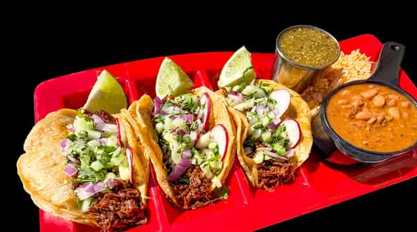 Three beef tacos with radishes, red onions, and lime wedges, with pinto beans, rice, and salsa on a red plate.