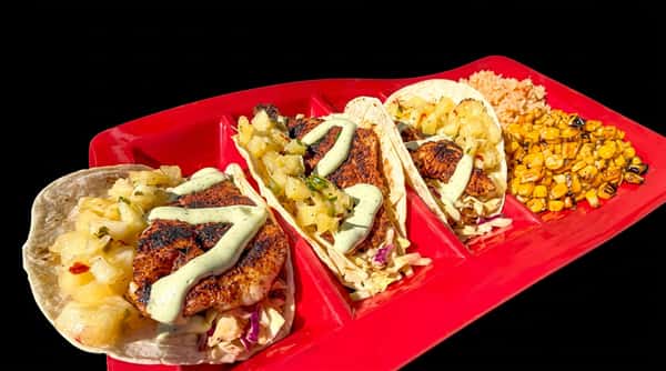 Three fish tacos with pineapple salsa and a creamy sauce, alongside rice and corn, on a red rectangular plate.