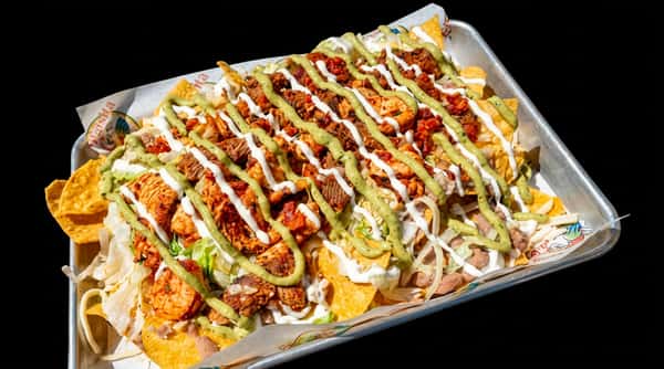 Nachos with chicken, beef, shrimp, lettuce, beans, and white & green sauces on a metal tray.