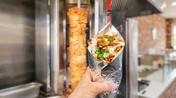Hand in a clear glove holding a shawarma wrap with chicken, white sauce, and green herbs, with a vertical meat spit in the background.