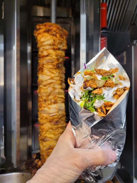 Fresh chicken döner wrap held in front of a traditional rotating shawarma spit with creamy sauce and herbs