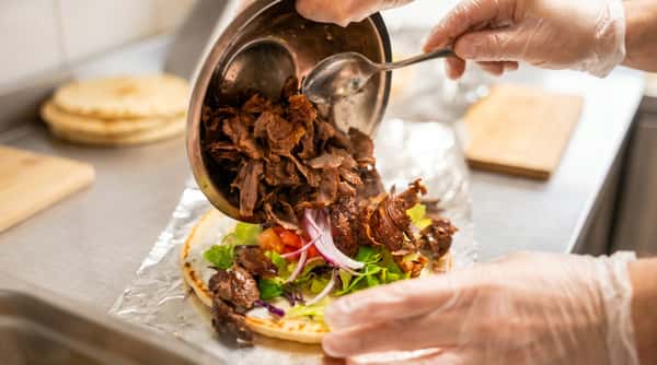 Gloved hands add shaved meat from a metal bowl onto a pita with lettuce, red onions, tomatoes, and white sauce.