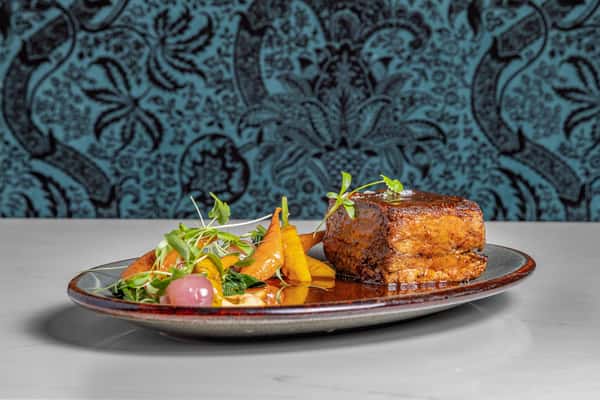 Slow-braised beef short rib glazed in rich sauce, plated alongside sautéed greens, heirloom carrots, and pickled onions, garnished with microgreens on a rustic ceramic plate.