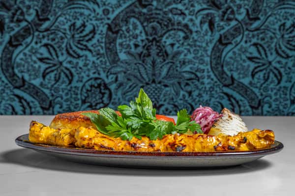 Juicy saffron chicken joojeh kebab served with golden tahdig, grilled tomato, pickled onions, and fresh herbs, plated on a patterned ceramic dish.