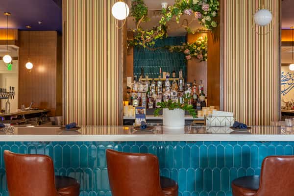 "Stylish Persian-inspired bar with turquoise tile facade, leather bar stools, a floral canopy, and a curated liquor display, creating a modern and luxurious dining atmosphere.