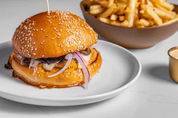 Gourmet chicken sandwich with grilled chicken breast, pickles, red onions, and creamy sauce on a toasted sesame brioche bun, served with a side of golden seasoned fries and dipping sauce on a white plate.