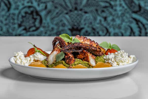 Charred Mediterranean-style grilled octopus served atop a bed of herbed green purée, garnished with roasted tomatoes, crumbled feta cheese, and fresh basil on a white plate with Persian-inspired backdrop.