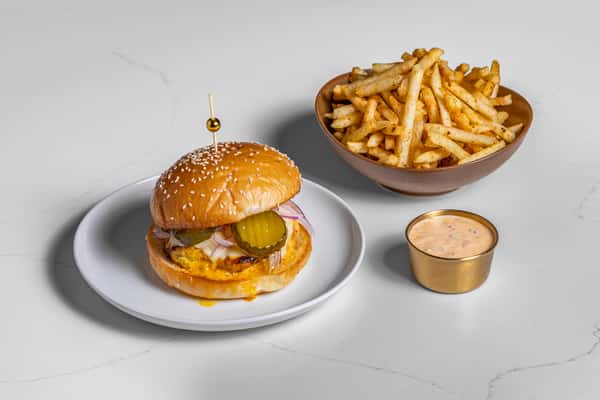 Halal grilled chicken burger with pickles, red onions, and melted cheese on a sesame bun, served with seasoned French fries and dipping sauce on the side.