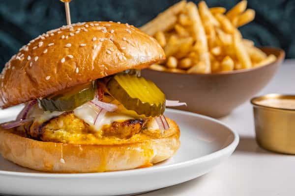 Juicy grilled chicken sandwich with pickles, red onions, and creamy sauce on a toasted sesame bun, served with golden French fries and a side of dipping sauce.