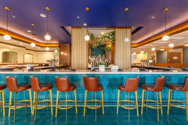Modern Persian restaurant bar with turquoise tiled counter, brown leather barstools, pendant globe lighting, and a backlit liquor display framed by wood paneling and floral accents.