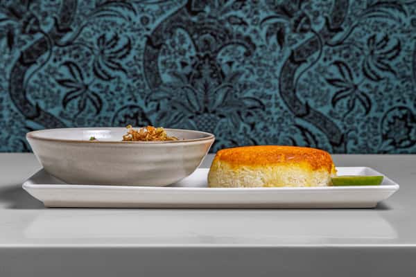 Traditional Persian soup paired with crispy saffron tahdig and a lime wedge, elegantly plated on a rectangular white tray against a Persian-patterned backdrop.