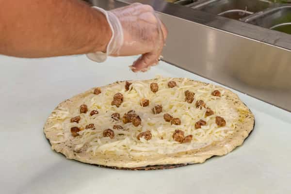 Unbaked white sauce pizza topped with shredded cheese and sausage crumbles, as a gloved hand adds final toppings at a prep station.