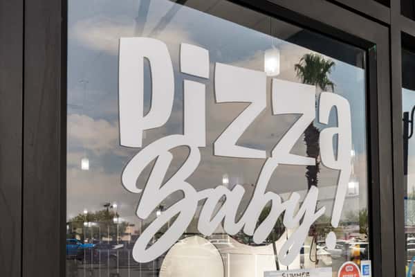 Large 'Pizza Baby!' logo decal on the front glass window of the restaurant, reflecting palm trees and a bright outdoor shopping plaza.