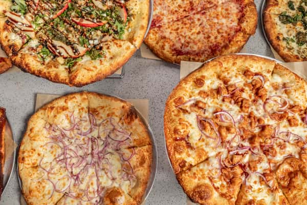 Assortment of artisan pizzas including BBQ chicken, cheese, veggie, white sauce, and gourmet options, displayed on a stone countertop.