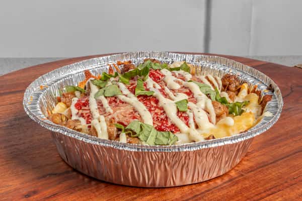 Baked pasta topped with Italian sausage, melted cheese, tomato sauce, fresh basil, and a drizzle of creamy dressing in a foil container.