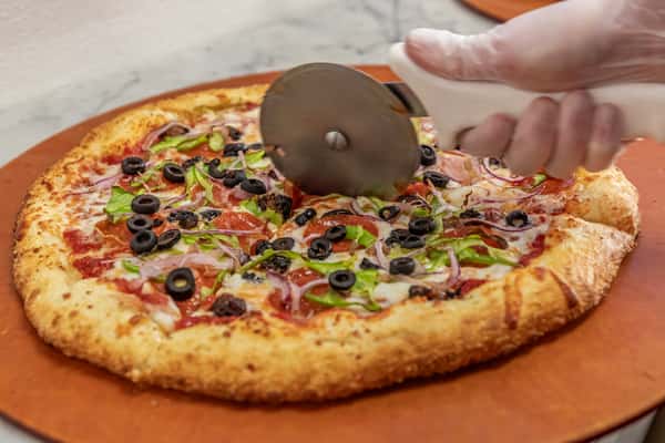 Freshly baked supreme pizza loaded with olives, red onions, green peppers, and pepperoni being sliced by hand in a pizzeria kitchen.