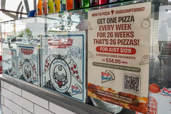 In-store display promoting Pizza Baby's pizza subscription deal, offering 26 pizzas for $129, with branded pizza boxes and colorful drinks in the background