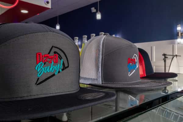 Close-up of branded 'Pizza Baby!' snapback hats in black and grey with embroidered logo, displayed on a glass counter inside a modern pizzeria.