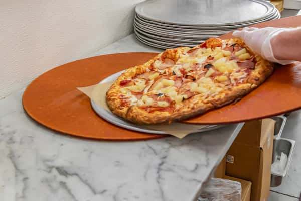 Freshly baked Hawaiian pizza with ham and pineapple being transferred to a serving tray in a busy pizzeria kitchen.