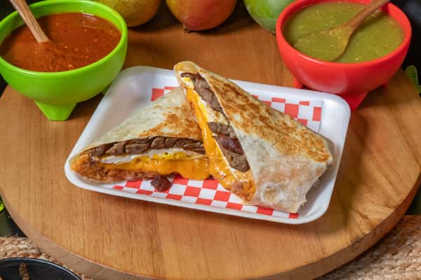 Grilled breakfast burrito filled with steak, melted cheddar cheese, eggs, and beans, cut in half to reveal a savory, melty interior. Presented on a checkered tray alongside green and red salsas, perfect for a hearty Mexican-style breakfast.